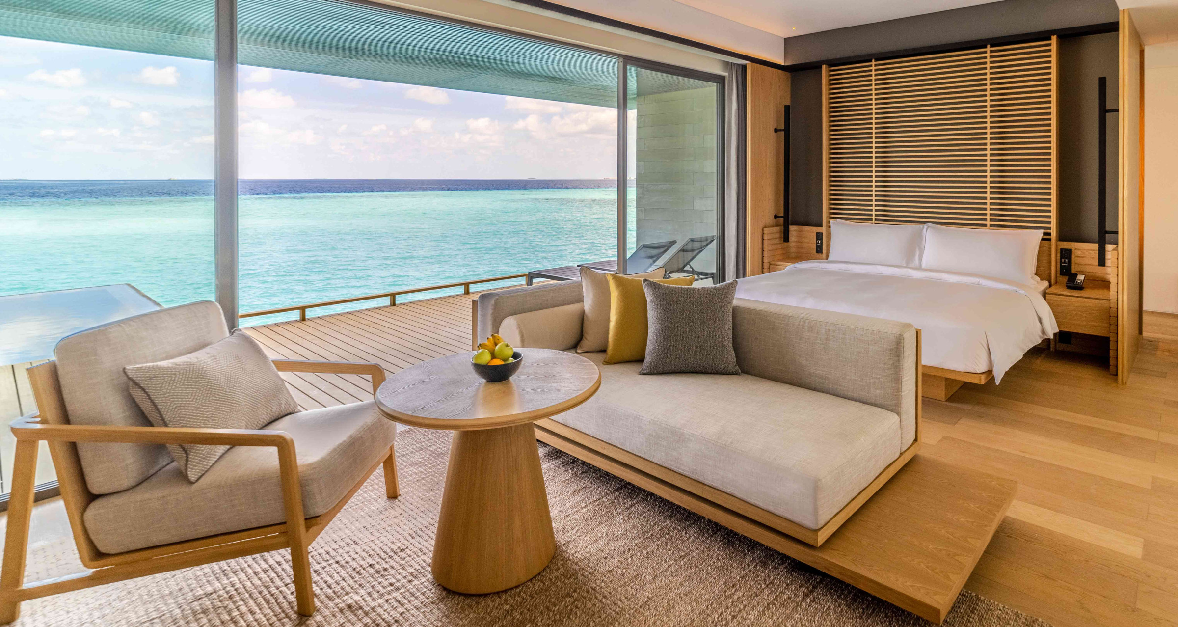 An image of a villa bedroom at Alila Kothaifaru Maldives, decorated in warm wooden tones and neutral furnishings. Large sliding doors open onto an outdoor deck that directly borders the ocean. 