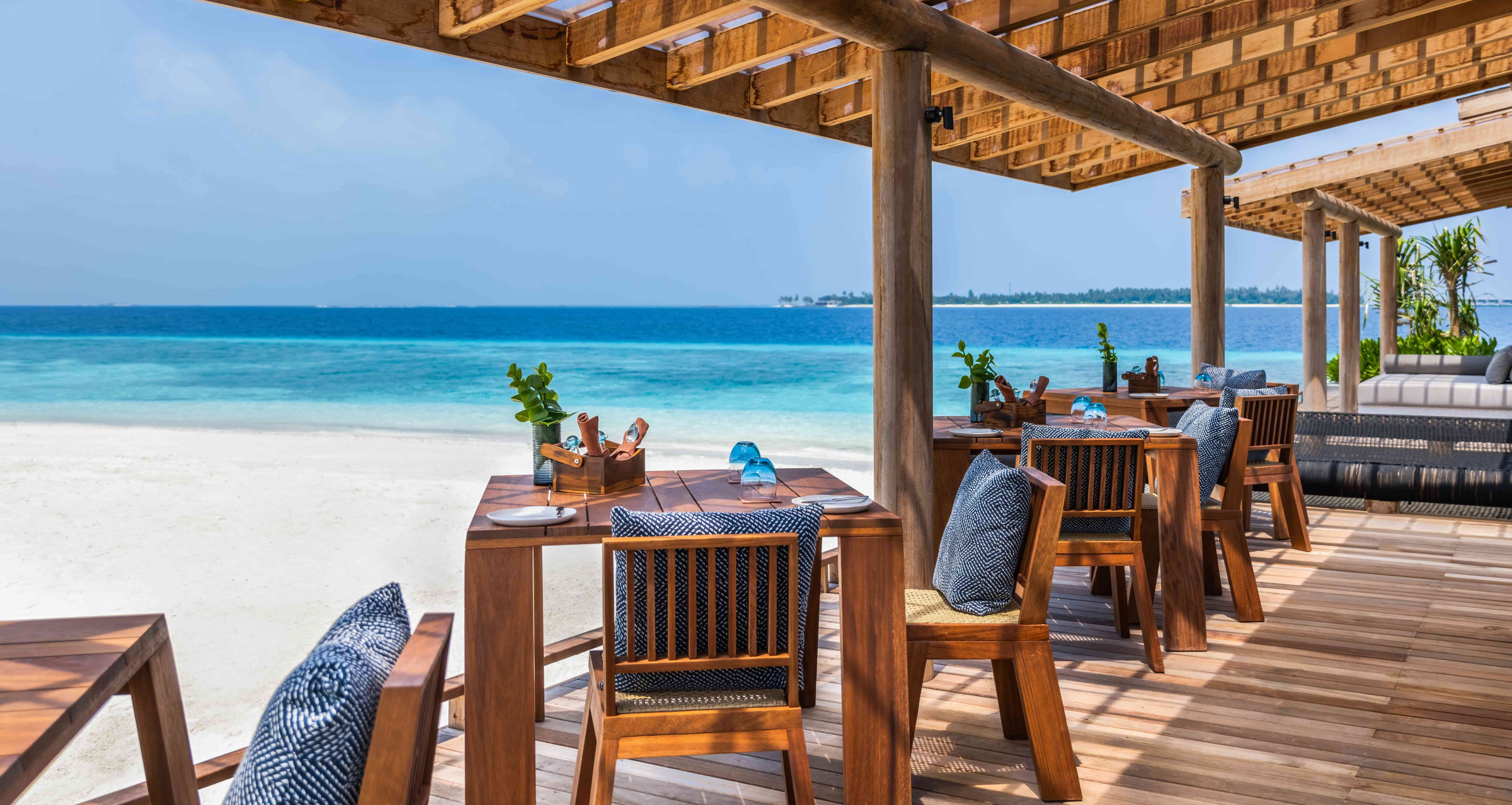 A view of a restaurant at Alila Kothaifaru Maldives. The restaurant is open sided and set on wooden decking, facing a white sand beach and the open ocean. 