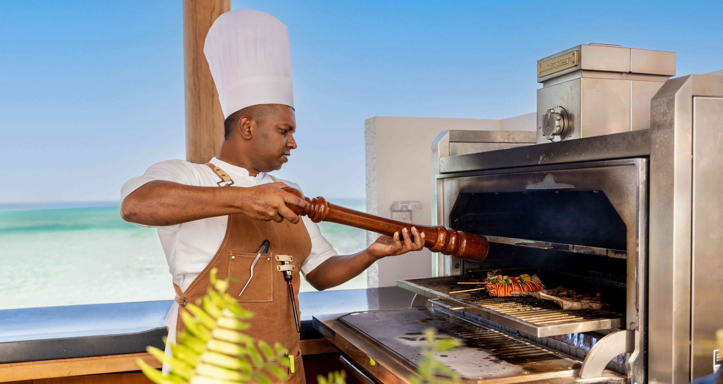 A chef at Alila Kothaifaru Maldives standing in front of a large oven, seasoning some food at is cooks. 