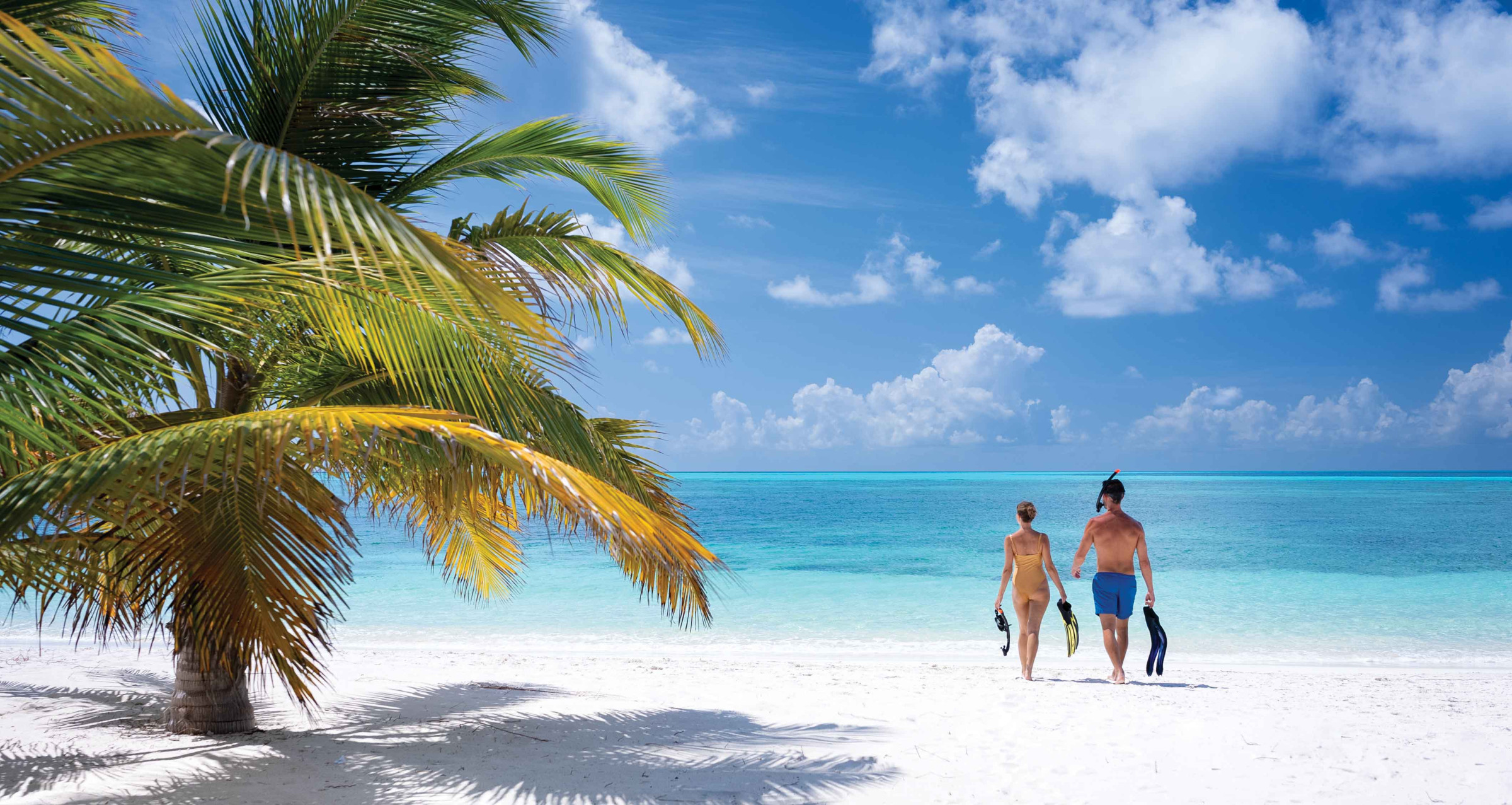 A male/female couple walking towards the ocean on a white sanded beach with snorkelling equipment in their hands.