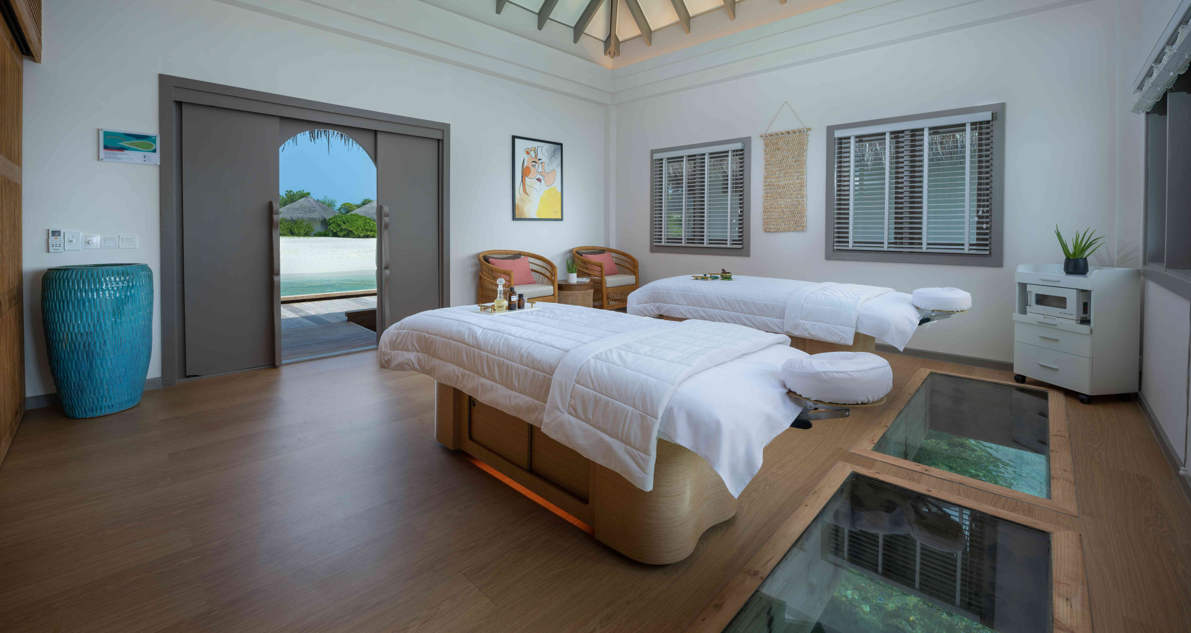 A view of an overwater couples' treatment suite at the MOSHKA Spa at Cora Cora Maldives. The two beds have head rests positioned over glass flooring that allows viewing of the sealife below while getting a treatment. 