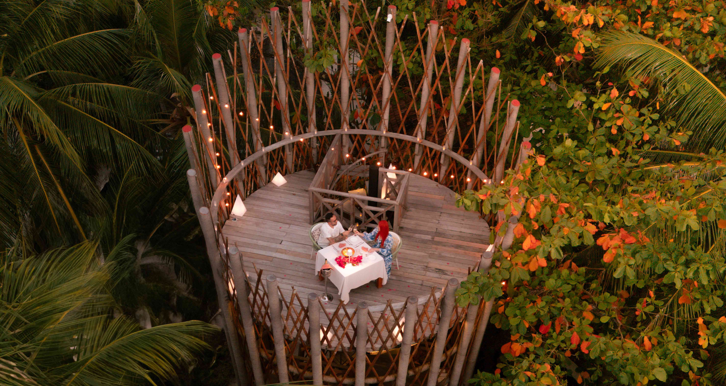 A couple dining atop a large and circular bamboo tower at Cora Cora Maldives. The treetop setting is framed by twinkling lights and the canopy.