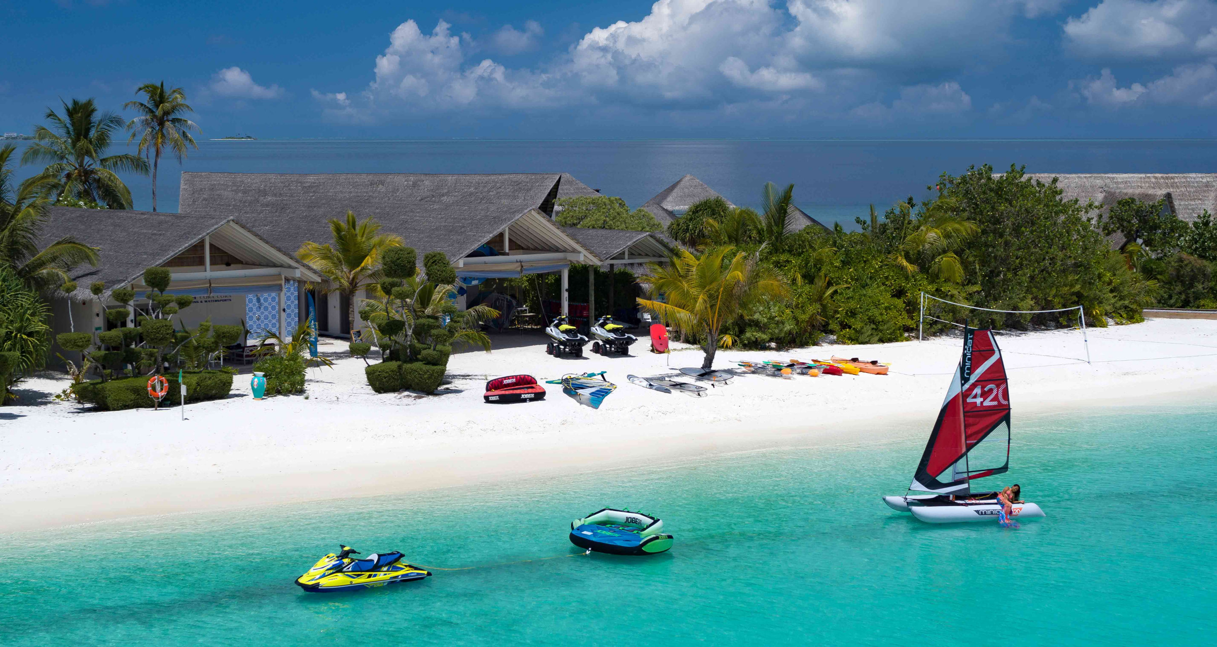 The water sports centre at Cora Cora Maldives, directly bordering the ocean on the beach. There are jet skis on the water and on the sand, as well as kayaks, paddle boards and parasails. 