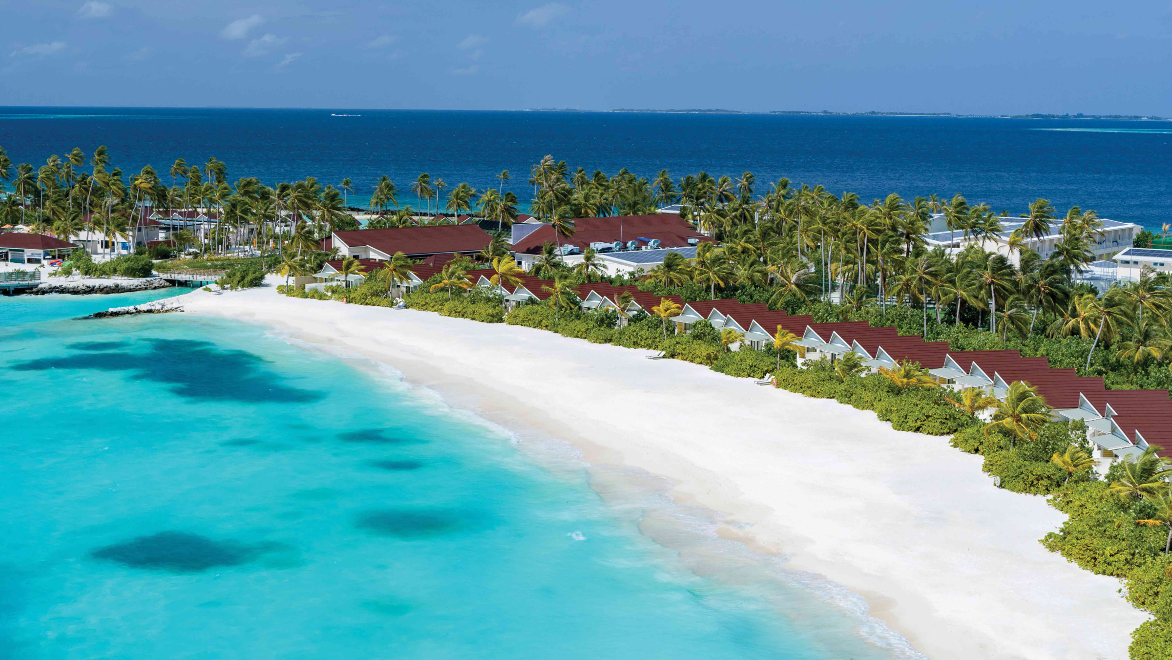 A view of the beach bordered by villas at OBLU XPERIENCE Ailafushi, a Colours of OBLU property, surrounded by green vegetation and facing the bright blue ocean. 