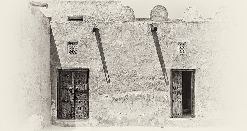 The History of Saadiyat Island - Sepia view of the inner courtyard of a traditional old Arabian adobe house in strong sunlight with shadows from wooden gutters and carved wooden doors and window lattices for ventilation.