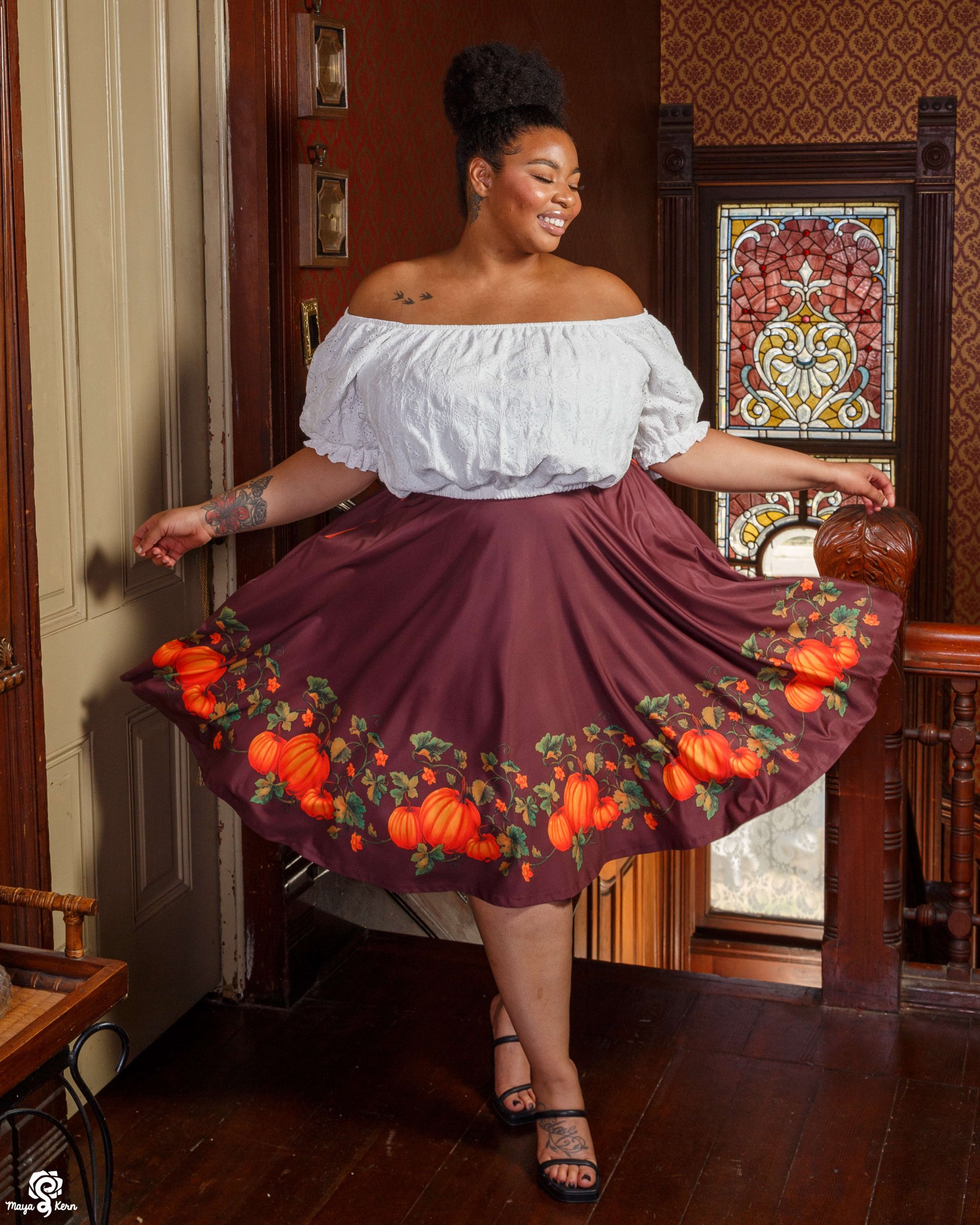 image of a plus size woman wearing a white eyelet crop top and a dark purple-ish brown midi skirt with a painting of orange pumpkins and green vines decorating the hem. it's the perfect fall fashion.