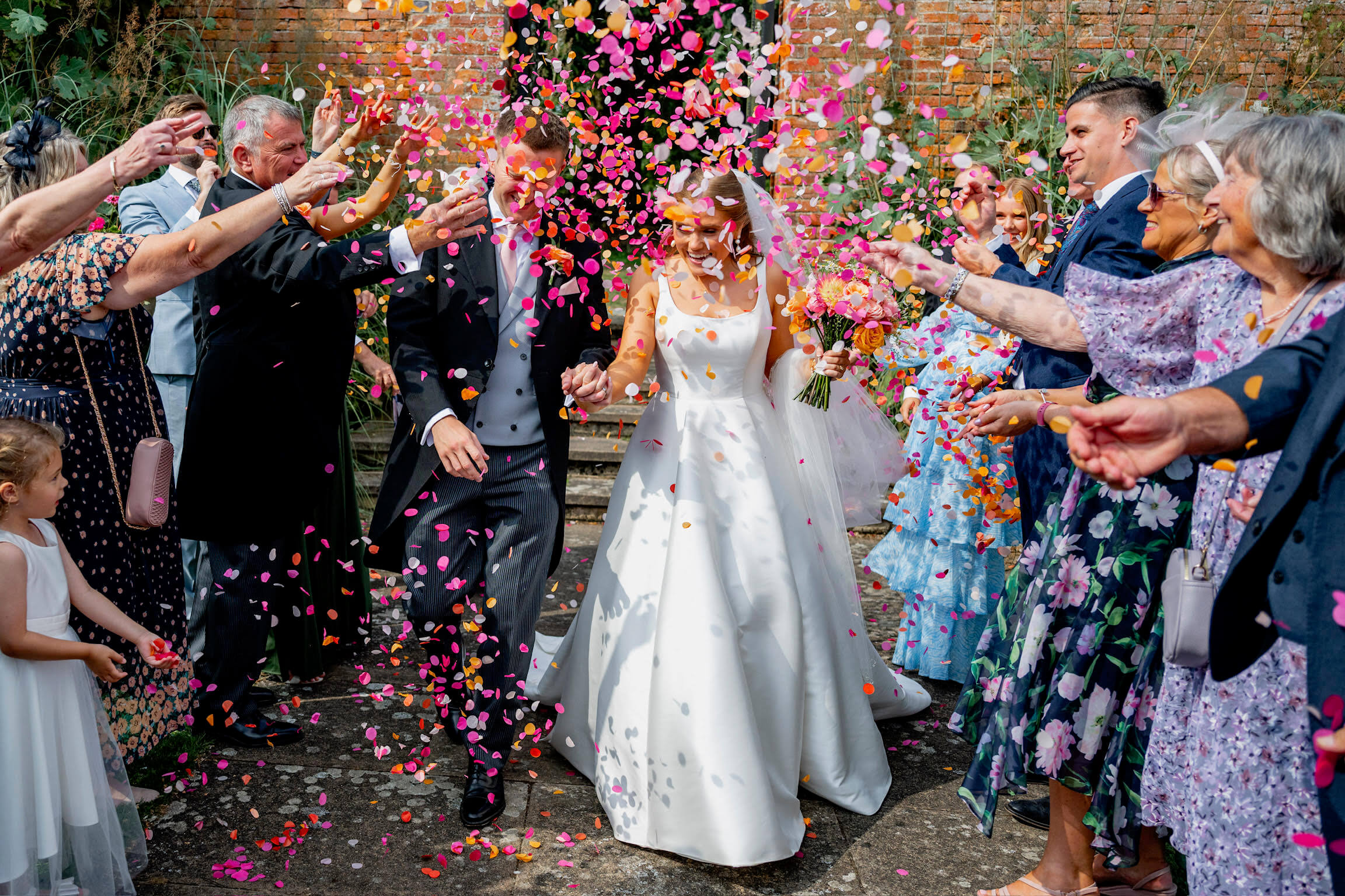 A groom and bride walk through a wedding confetti storm of pinks and oranges. Their guests are smiling as they throw the wedding confetti.