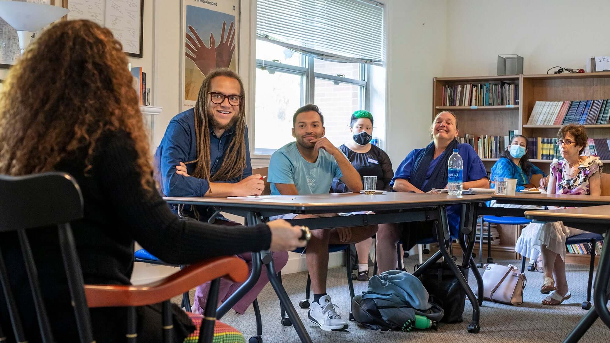 A group of people seated around tables in a classroom or meeting room, engaged in a discussion. Bookshelves line the back wall, and natural light comes in through a large window. A person in the foreground faces the group while speaking or presenting.