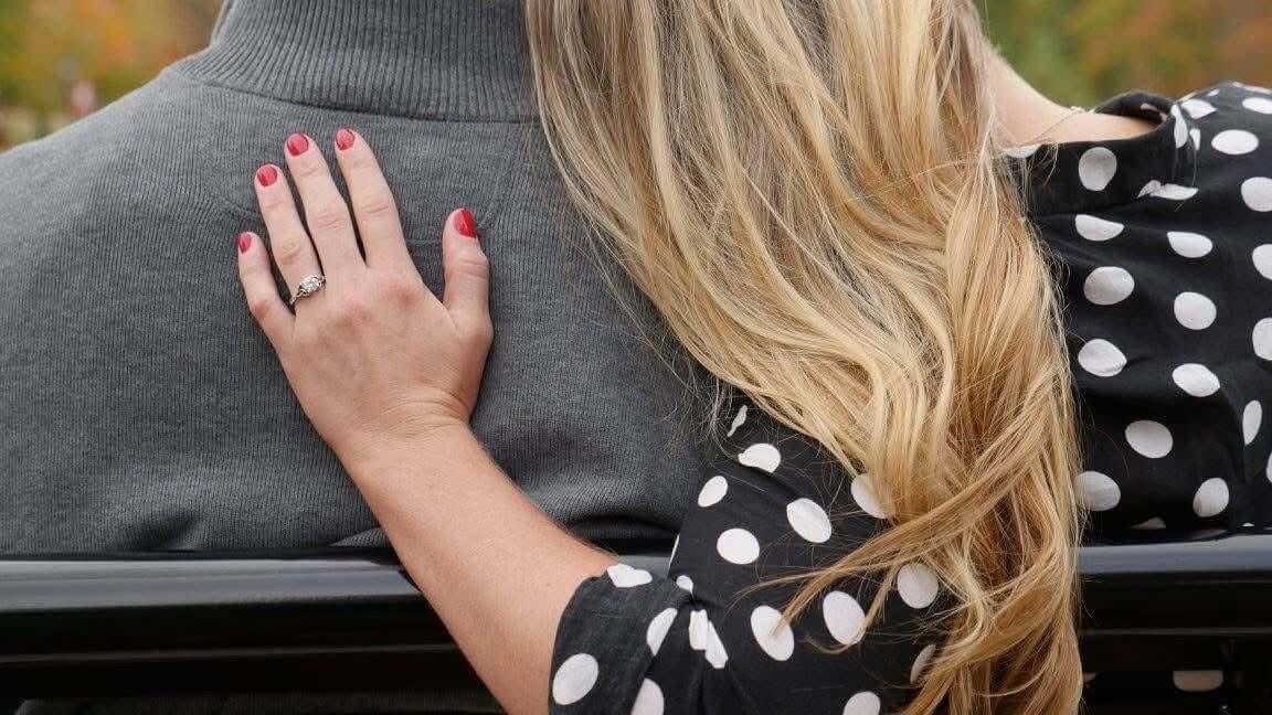 Two people sitting on a park bench with arms around each other, looking out at autumn scenery.