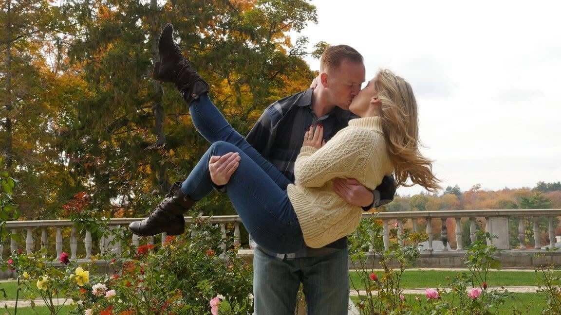 A person lifting another in a garden area surrounded by flowers and autumn trees.
