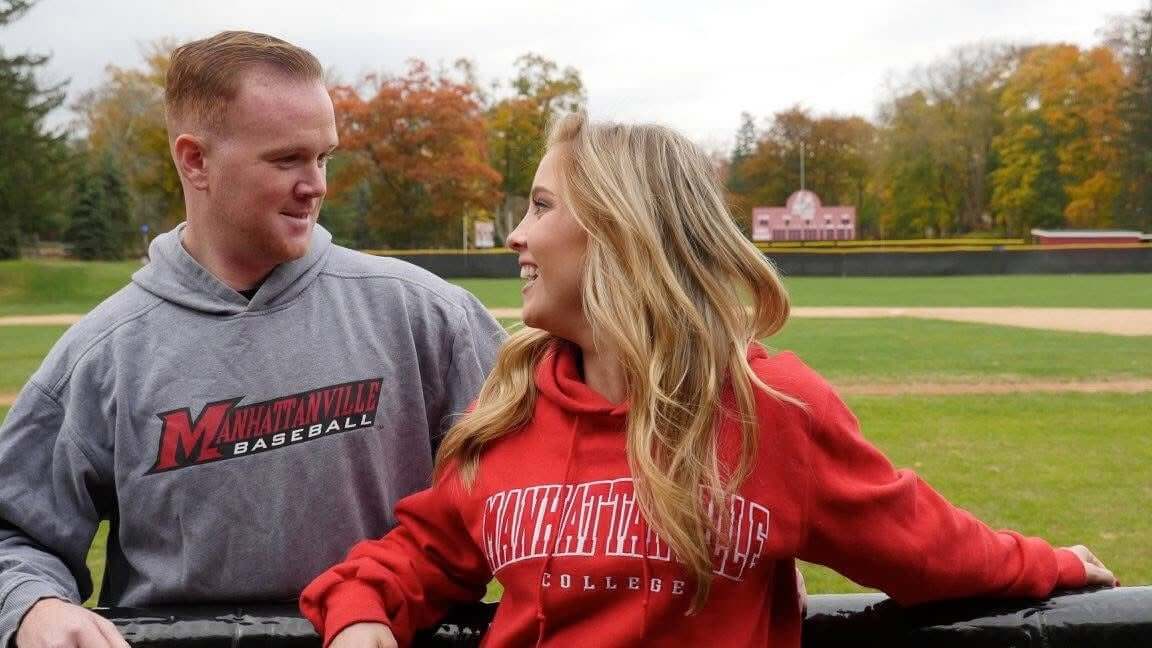 wo people standing at a baseball field railing wearing matching college‑branded sweatshirts.