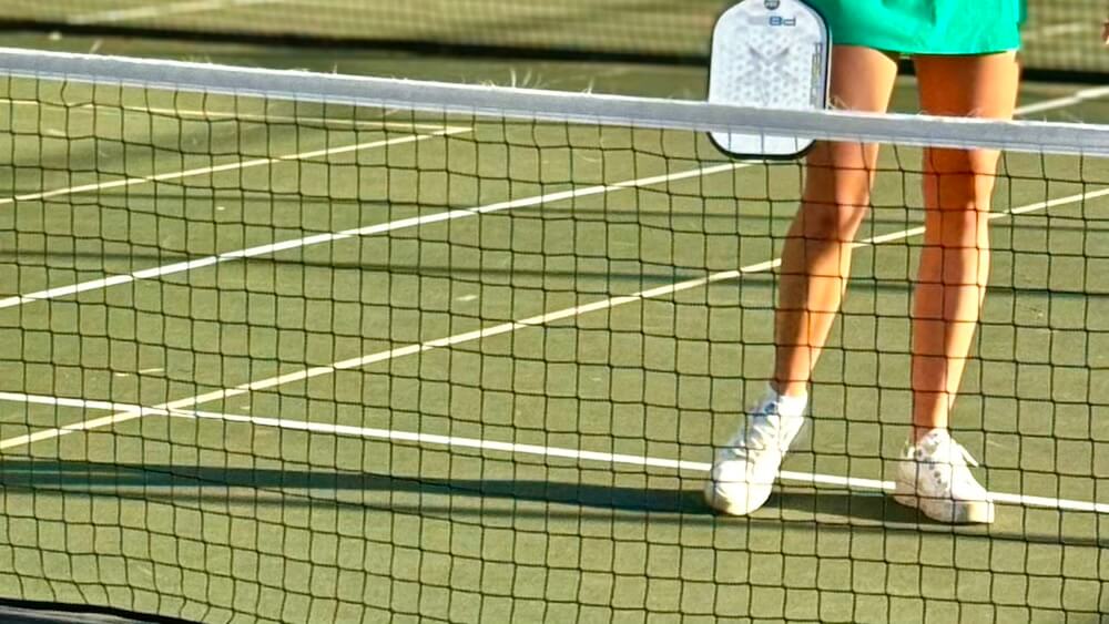 Close up on a pickleball net and one player's legs and a green court