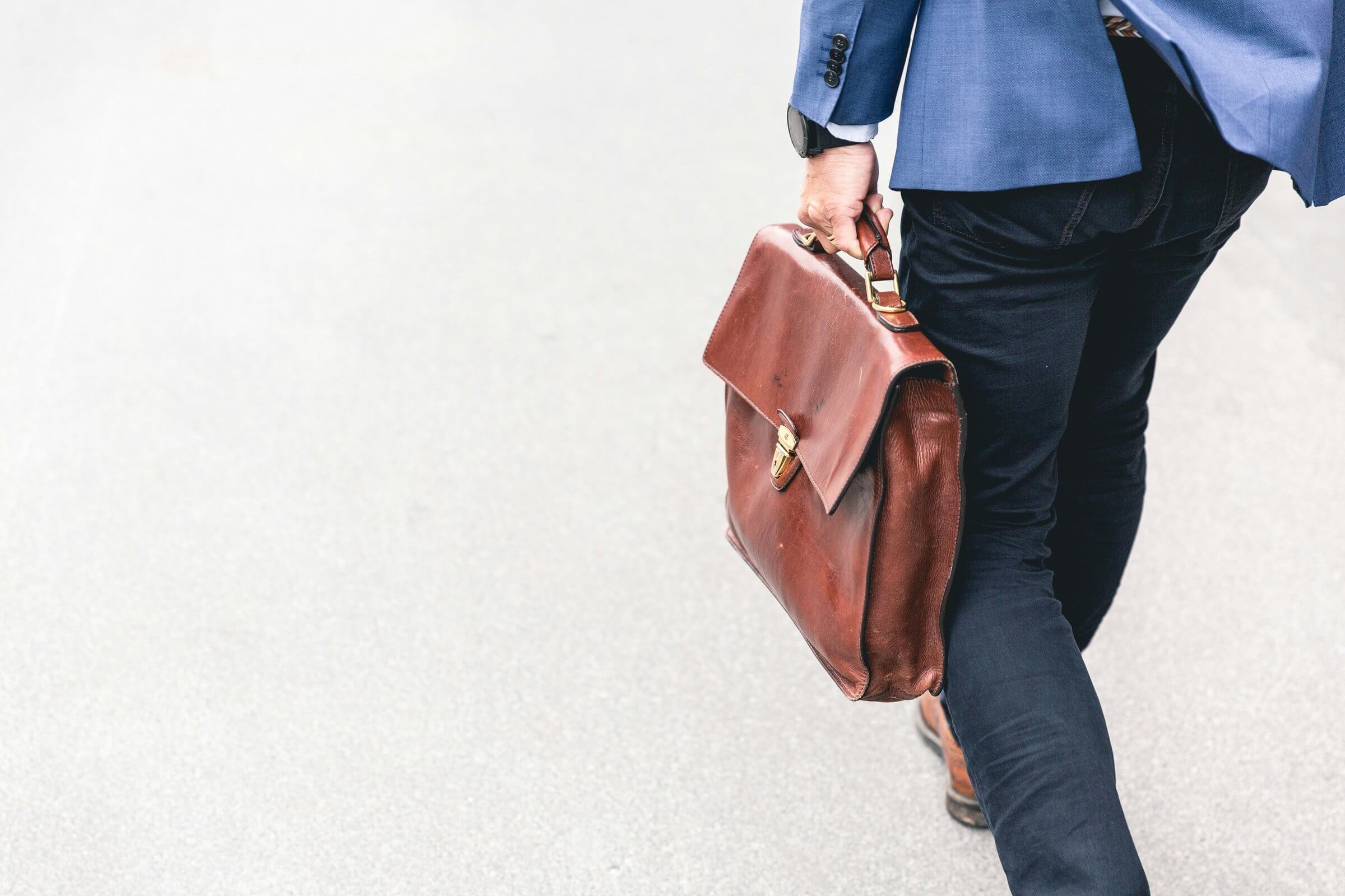 man in dark jeans and blue blazer walking away holding leather bag in left hand