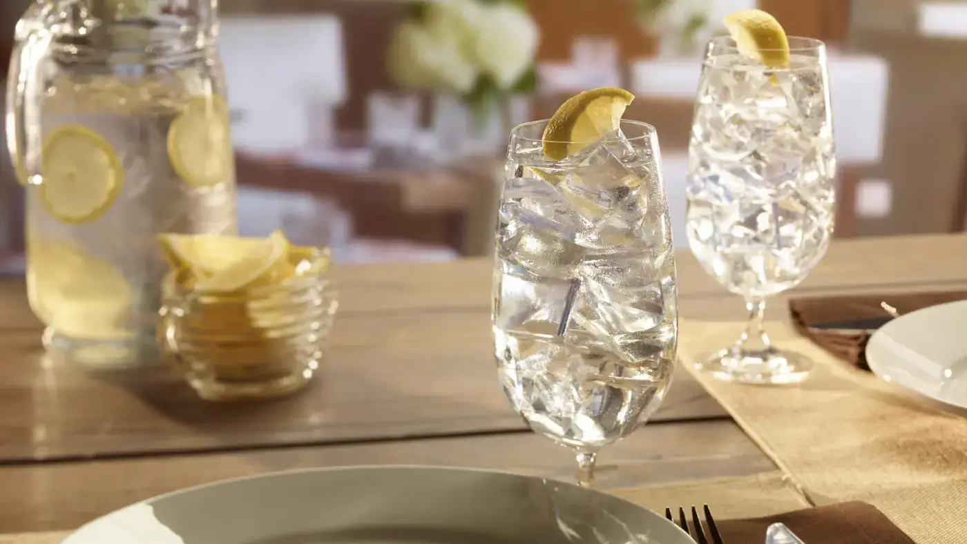 A close-up of two tall glasses filled with ice cubes and water on a dining table. The glasses have a lemon slice garnish. A pitcher filled with water and lemon slices is visible in the background. The table is set with plates and cutlery.