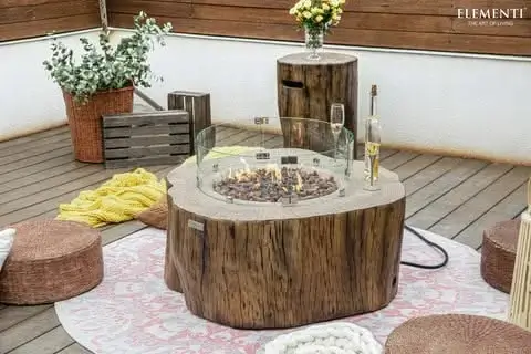Elementi Manchester Concrete Gas Fire Pit Table, designed to look like a tree stump with a glass wind guard and lit flames over fire rocks, on an outdoor patio with matching tree stump-style tank cover and seating.