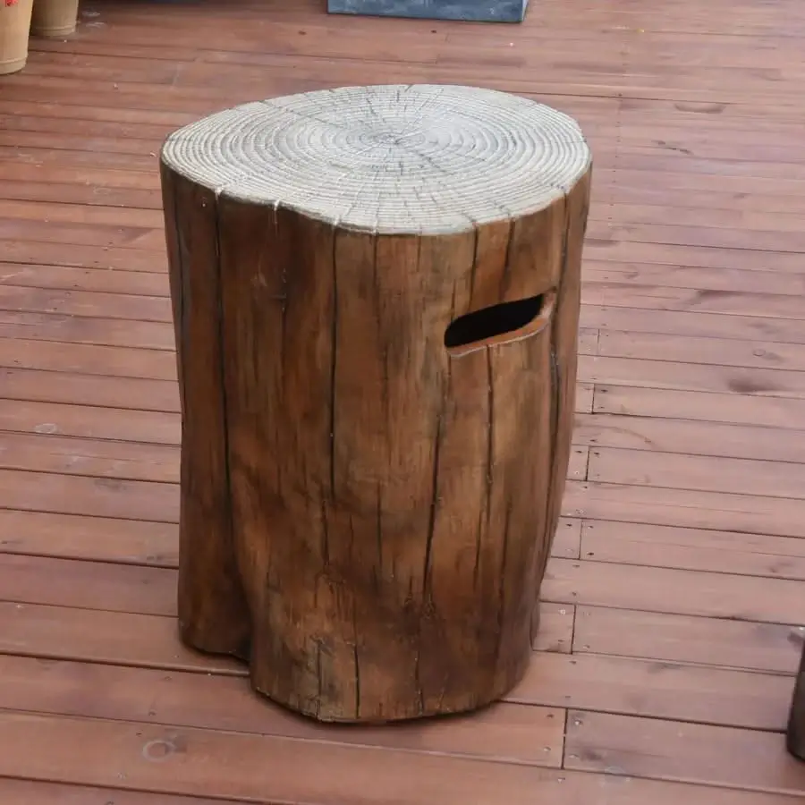 A propane tank cover resembling a tree stump, possibly the Elementi Manchester Redwood Propane Tank Cover, with a wood grain texture and handle cutout, shown on a wooden deck.