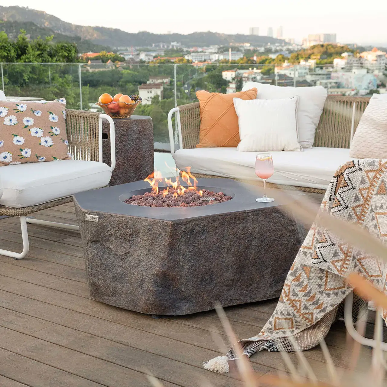 Elementi Columbia Outdoor Fire Pit Table, a concrete propane gas fire pit table with a stone-like texture and lit flames, on a rooftop patio with seating, pillows, and a city view.