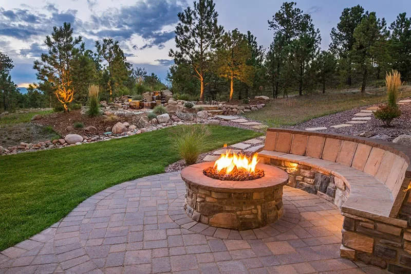 A circular stone fire pit with a glowing flame, surrounded by a curved stone bench. The backyard features a landscaped area with trees, shrubs, and a winding stone pathway.