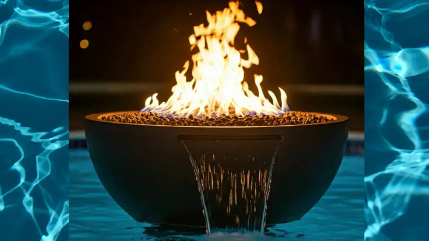 A sleek, modern concrete fire and water bowl sits partially submerged in a swimming pool at night. The bowl is filled with glowing embers and a roaring fire, while a gentle waterfall cascades over its edge into the water. The scene is illuminated by the warm glow of the fire.