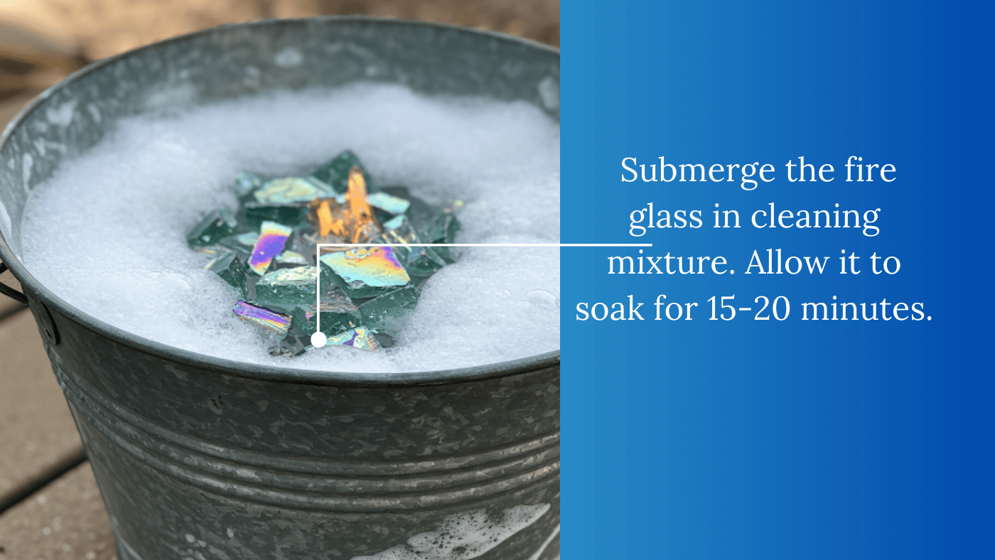 A close-up of a galvanized metal bucket filled with a soapy water solution. The solution is slightly foamy, and multicolored fire pit glass is submerged within it. The text "Submerge the fire glass in cleaning mixture. Allow it to soak for 15-20 minutes." is overlaid on the image.