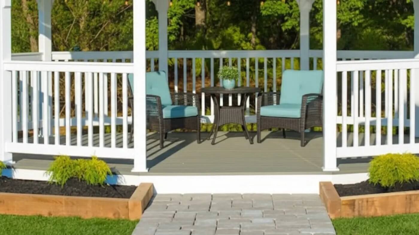 A pristine white vinyl gazebo featuring a modern gray composite floor, furnished with two comfortable wicker chairs and a small table. Lush green landscaping with small yellow shrubs borders the gazebo, and a paver walkway leads to its entrance.