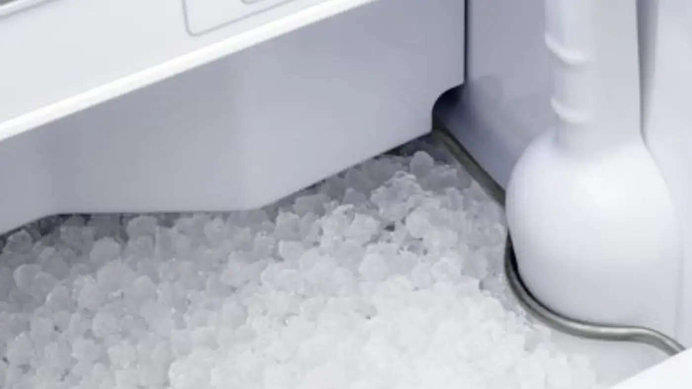 A close-up view of an ice maker bin. The bin is filled with a large quantity of crescent-shaped ice cubes. A white plastic scoop is visible in the bin, used to dispense the ice. The ice cubes are glistening and appear to be freshly made.