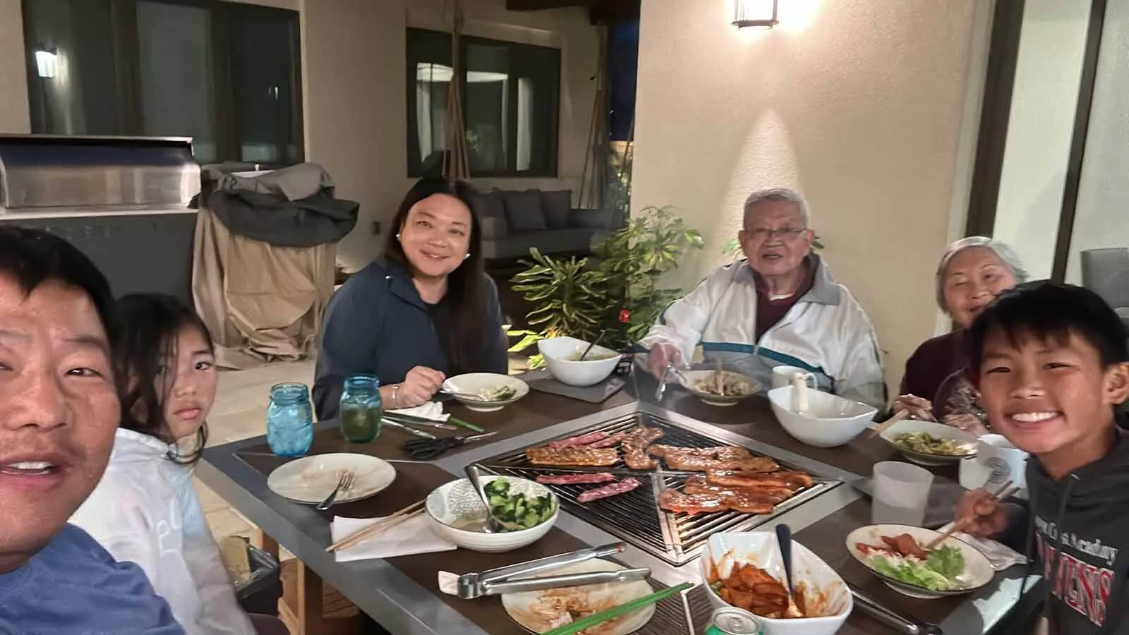  A multigenerational Asian family is gathered around a bbq grill table for an evening meal. The family is sitting closely together, smiling at the camera as they eat. On the grill in the center of the table, various meats and sausages are cooking.