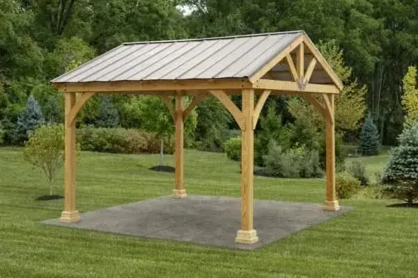 A light-colored wooden a frame pavilion with a gray metal roof, resting on a square concrete pad in a green, grassy outdoor setting.