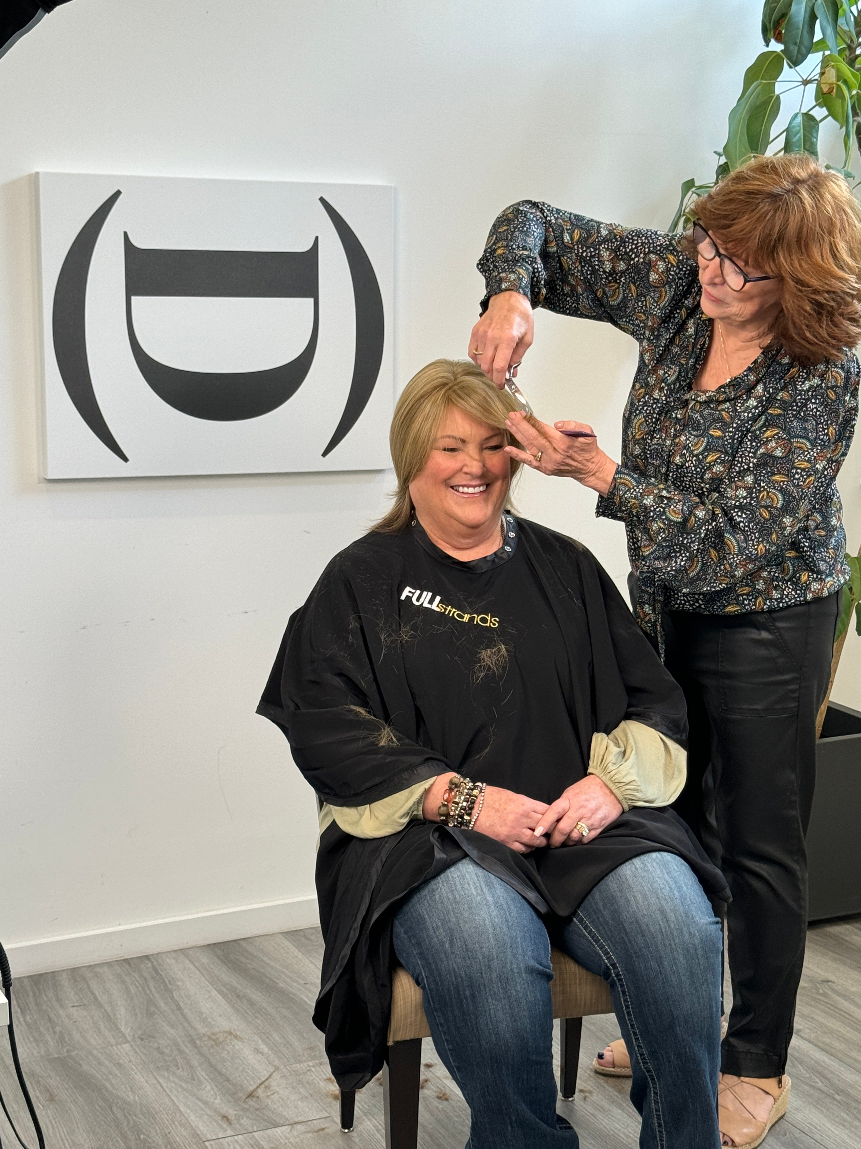 A stylist assists a woman in fitting a human hair topper, highlighting a collaborative approach to hair styling.