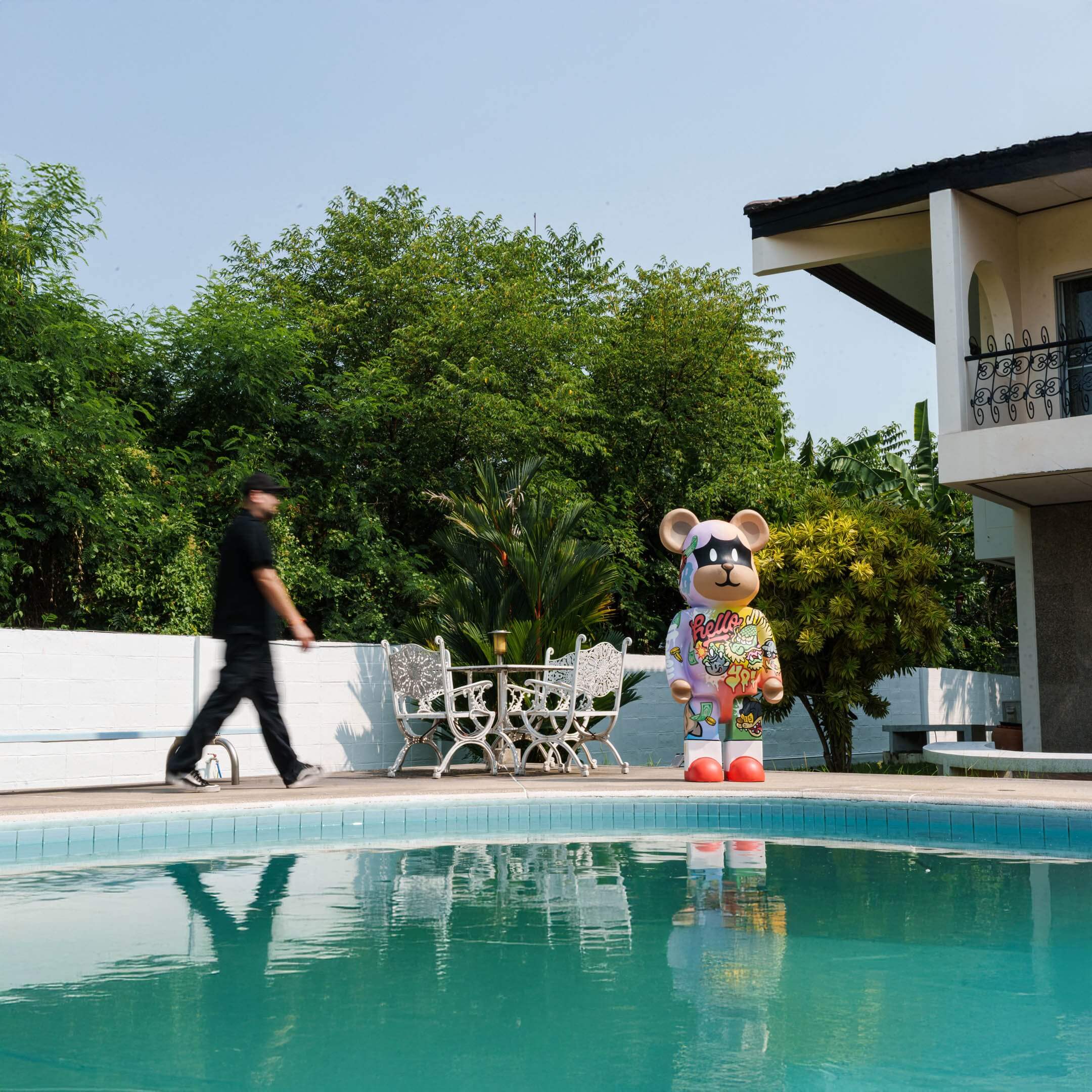 Lucky Paws street-art bear statue stands poolside beside an ornate white patio set, with lush greenery, a modern villa, and turquoise water completing the scene.