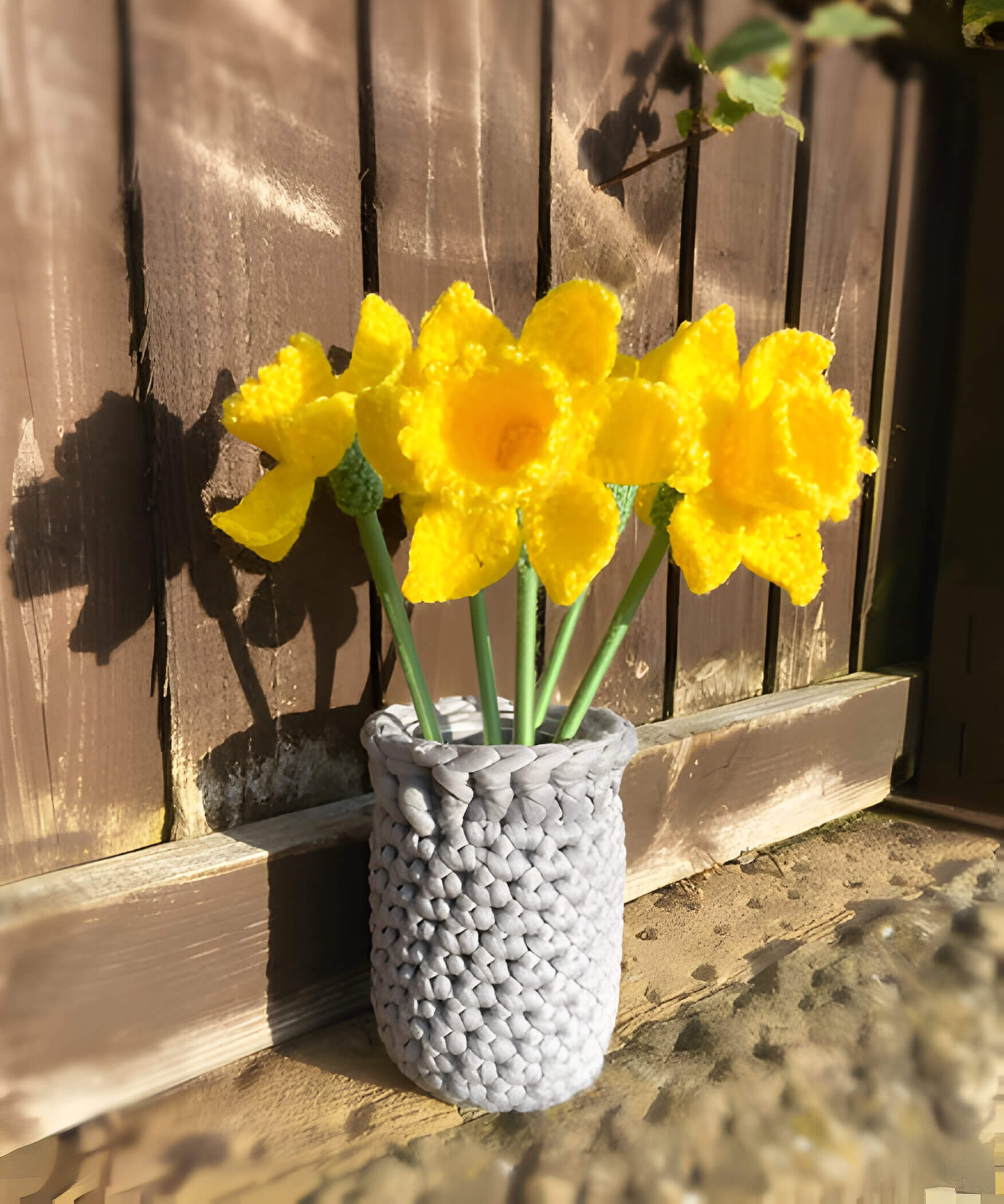 a crochet grey vase with yellow crochet daffodils against a brown garden fence
