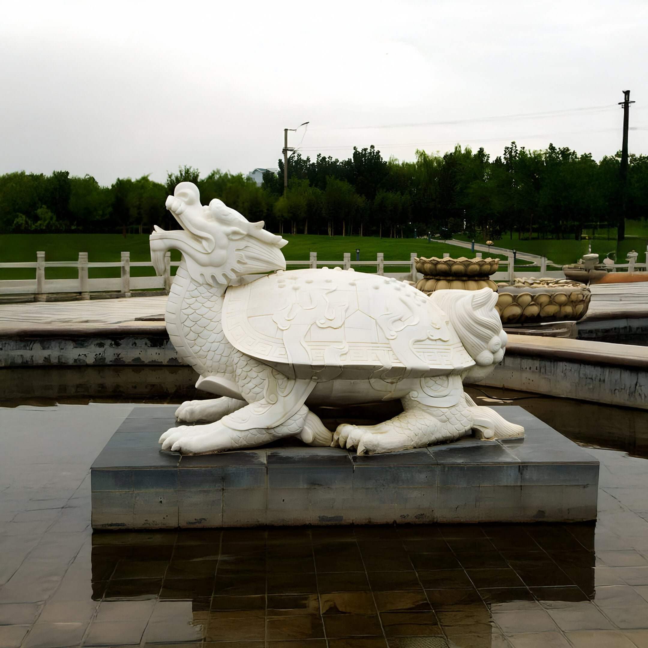 The Chinese Guardian Dragon Marble Outdoor Sculpture by Giant Sculptures (80cm), a hand-carved white marble piece blending a turtle with a dragons head, is elegantly displayed on a platform in shallow water amidst pathways, lush greenery, and an overcast sky.