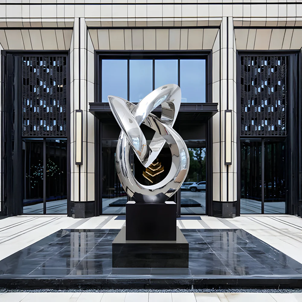 Large polished silver abstract looped sculpture on a black pedestal in front of a modern glass-and-tile building entrance, reflecting the surrounding plaza and sky.