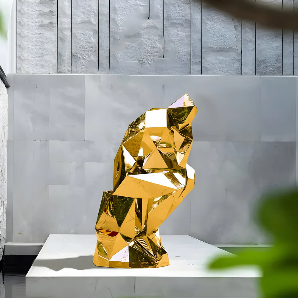 Large gold faceted abstract sculpture on a white outdoor platform, set against a light gray textured wall, with sharp geometric surfaces reflecting light and a blurred green plant in the foreground.