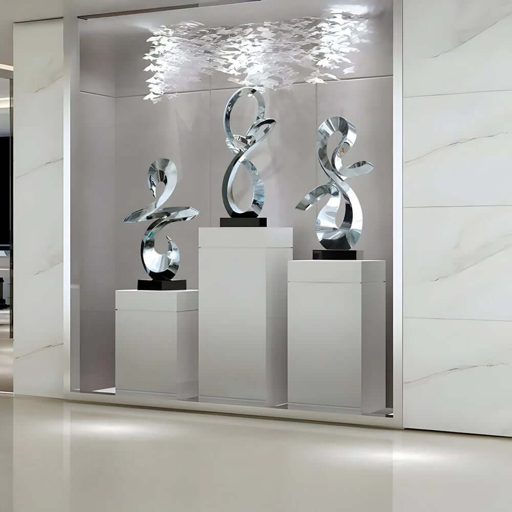 Three tall silver abstract ribbon sculptures on white pedestals in a recessed, modern display nook with soft overhead lighting and a cluster of delicate white hanging elements above.