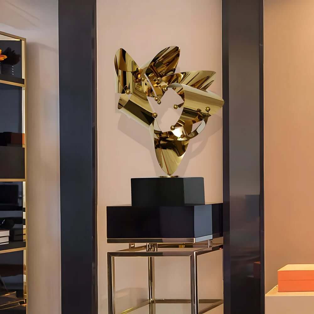 Polished silver abstract sculpture with sharp, wing-like extensions displayed on a pedestal in a modern gallery setting.