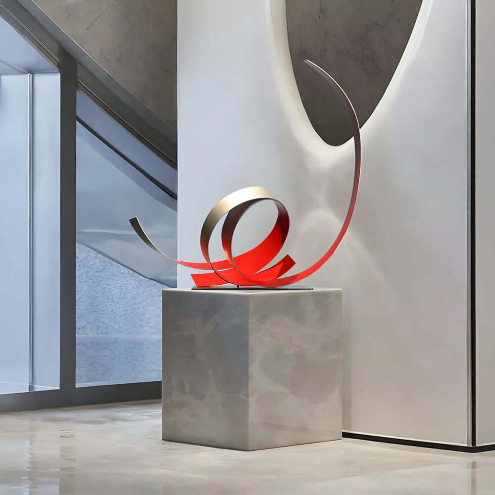 Modern abstract sculpture with red and gold metallic ribbons displayed on a marble pedestal in a contemporary indoor setting.