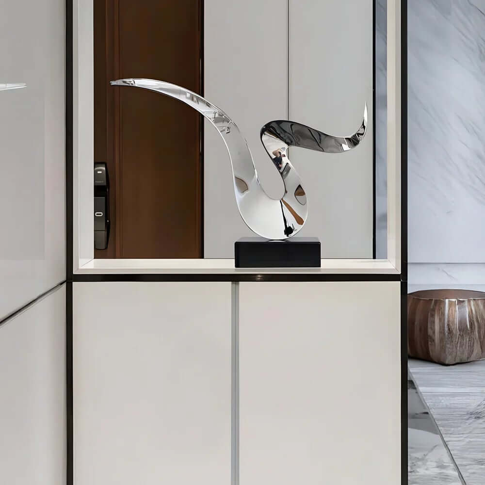 Sleek abstract metal sculpture with smooth, flowing curves displayed on a black base in a modern minimalist interior.