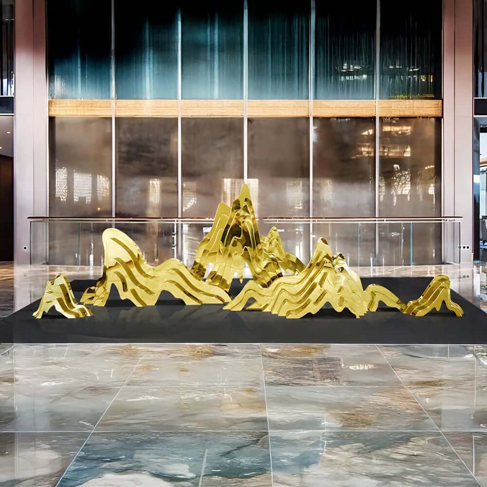 Large gold mirror-finish abstract “mountain range” sculpture installed in a modern lobby with glass façade and reflective marble floor.