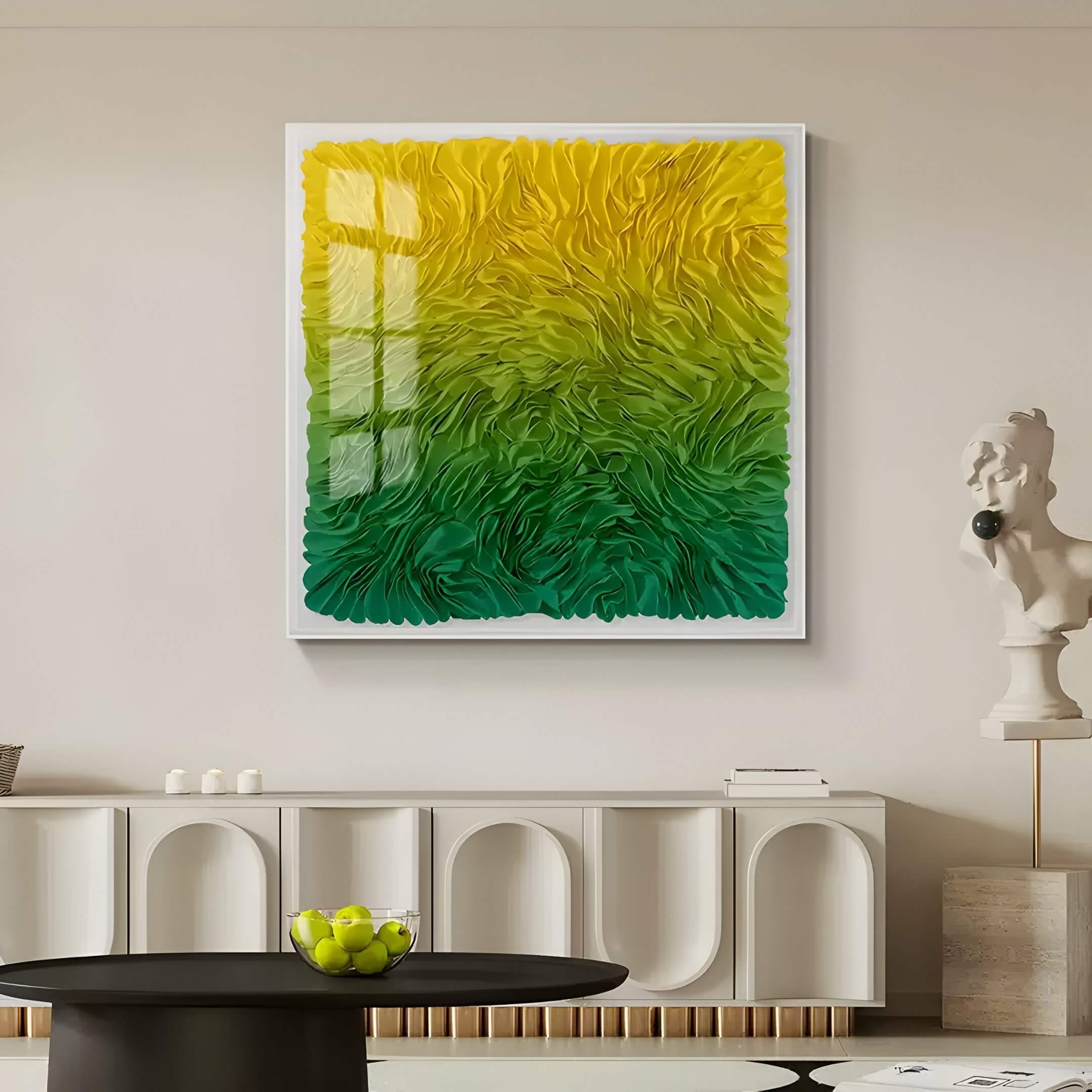 Square framed 3D wall art with layered ribbon textures in a yellow-to-green gradient, displayed above a modern white sideboard in a neutral-toned living room.