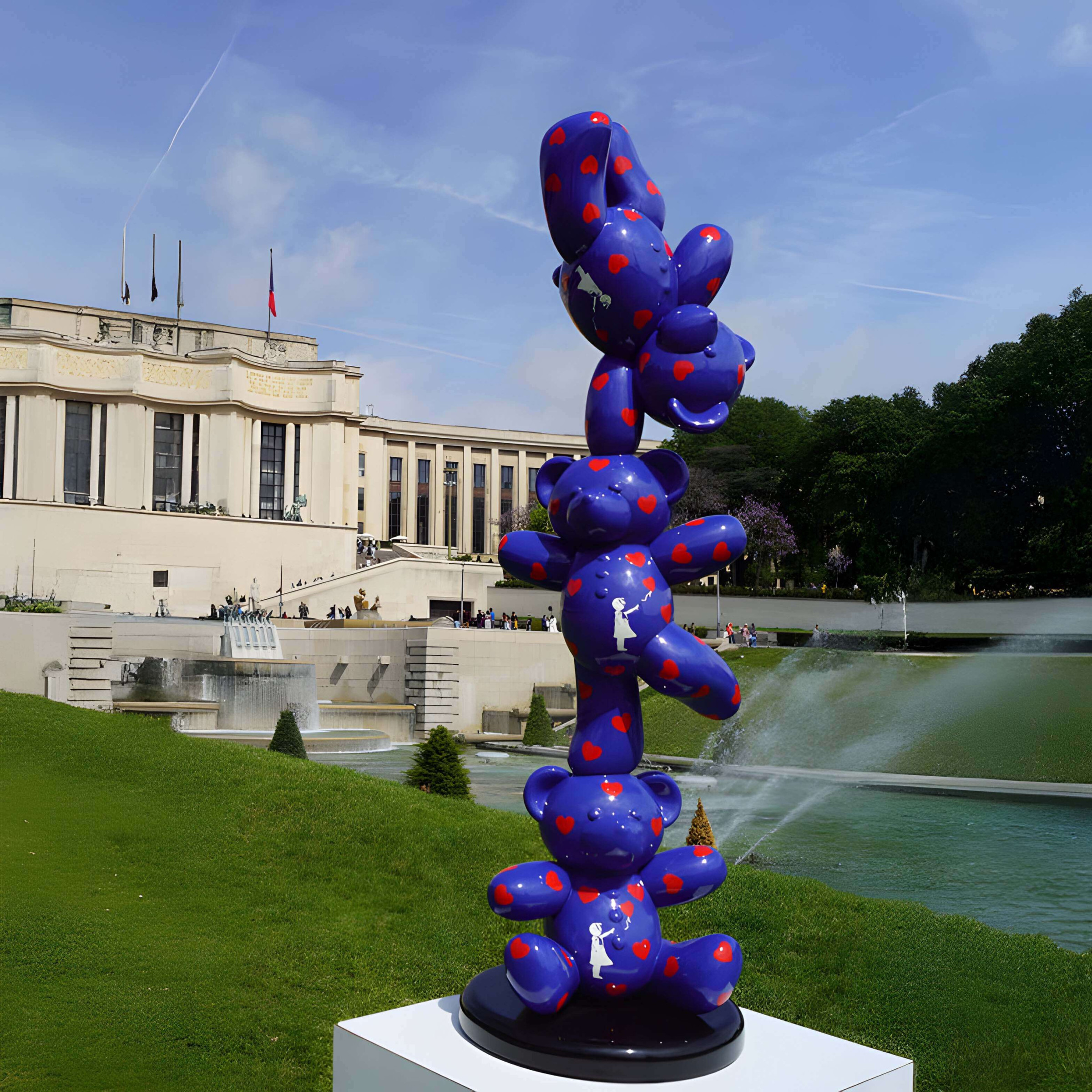Stacked Blue Bear Sculpture – A vibrant blue bear sculpture tower with red hearts, set in a scenic park with fountains and a grand building.