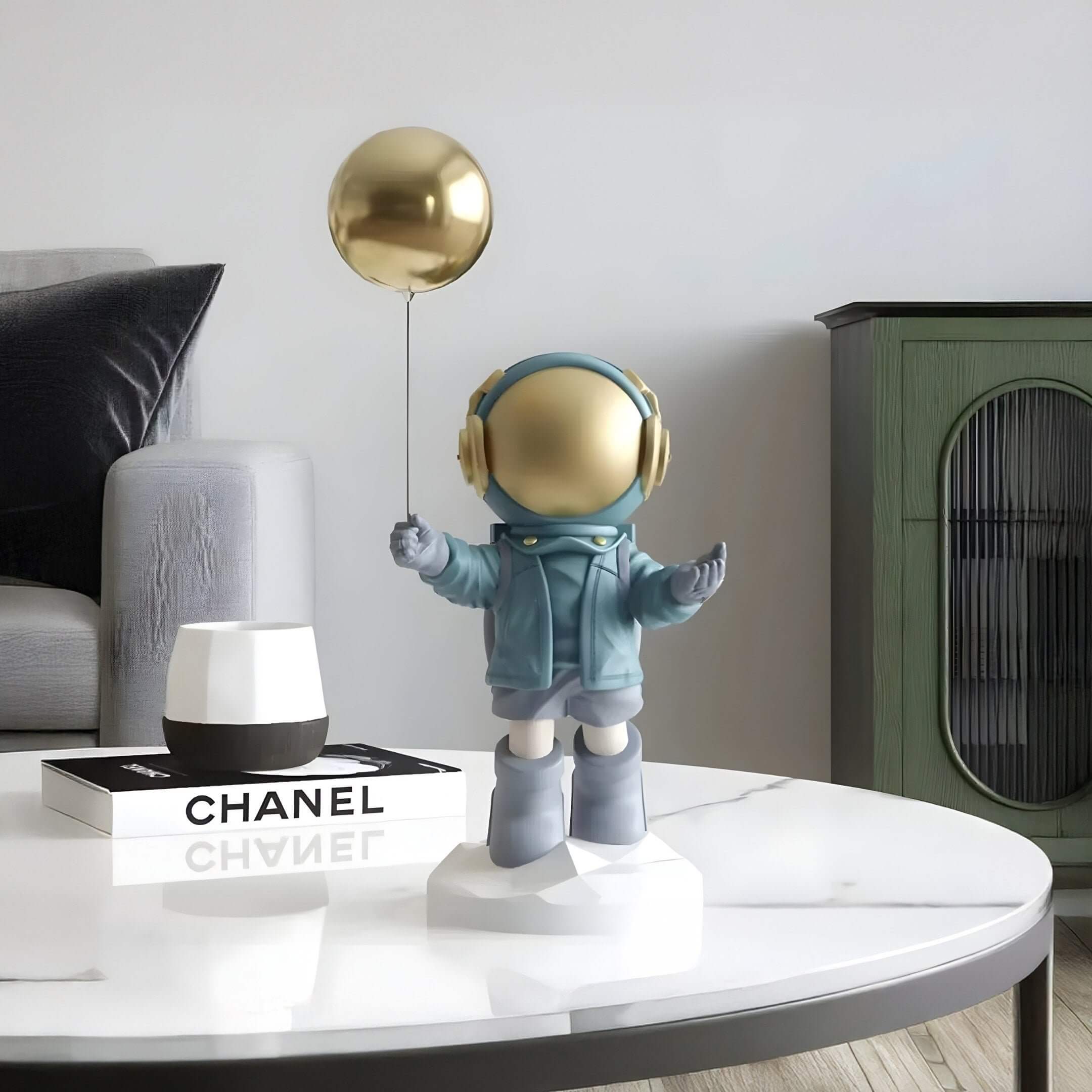 Blue astronaut sculpture with a gold visor holding a gold balloon on a geometric base, displayed on a white marble coffee table in a modern living room.