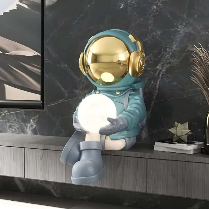Decorative astronaut figurine in a blue suit with a golden helmet holding a glowing orb, sitting on a shelf against a dark marble wall.