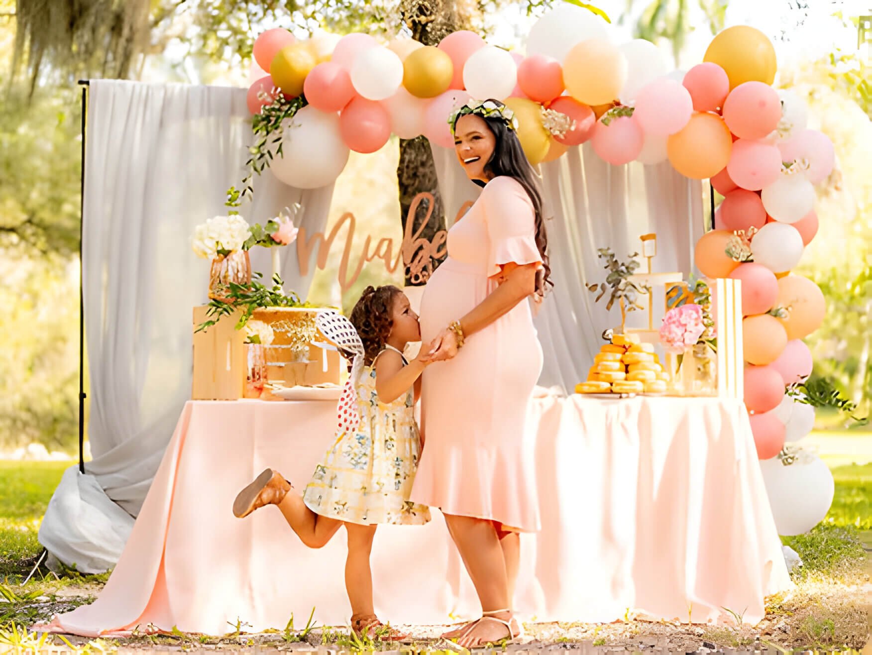 A woman at a baby shower with decorations behind, and child kissing her bump