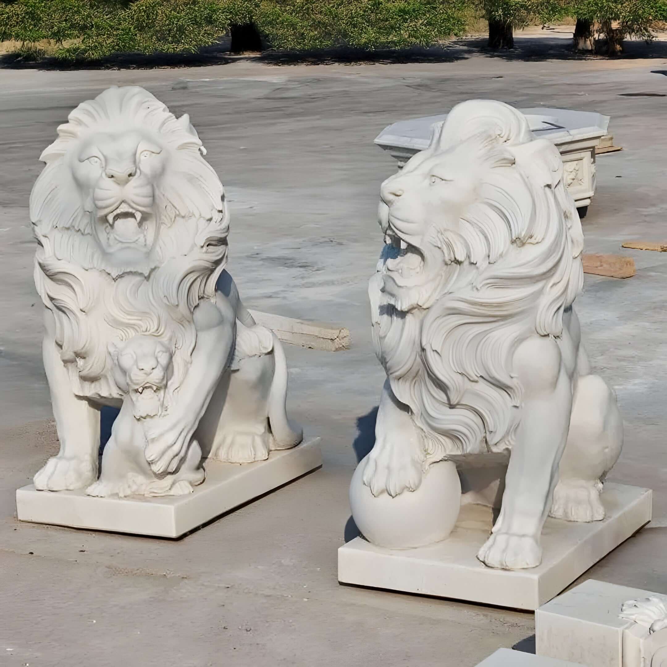 White stone lion custom animal sculptures featuring intricate detailing, one with a cub and the other with a paw resting on a sphere, displayed outdoors on a concrete surface.