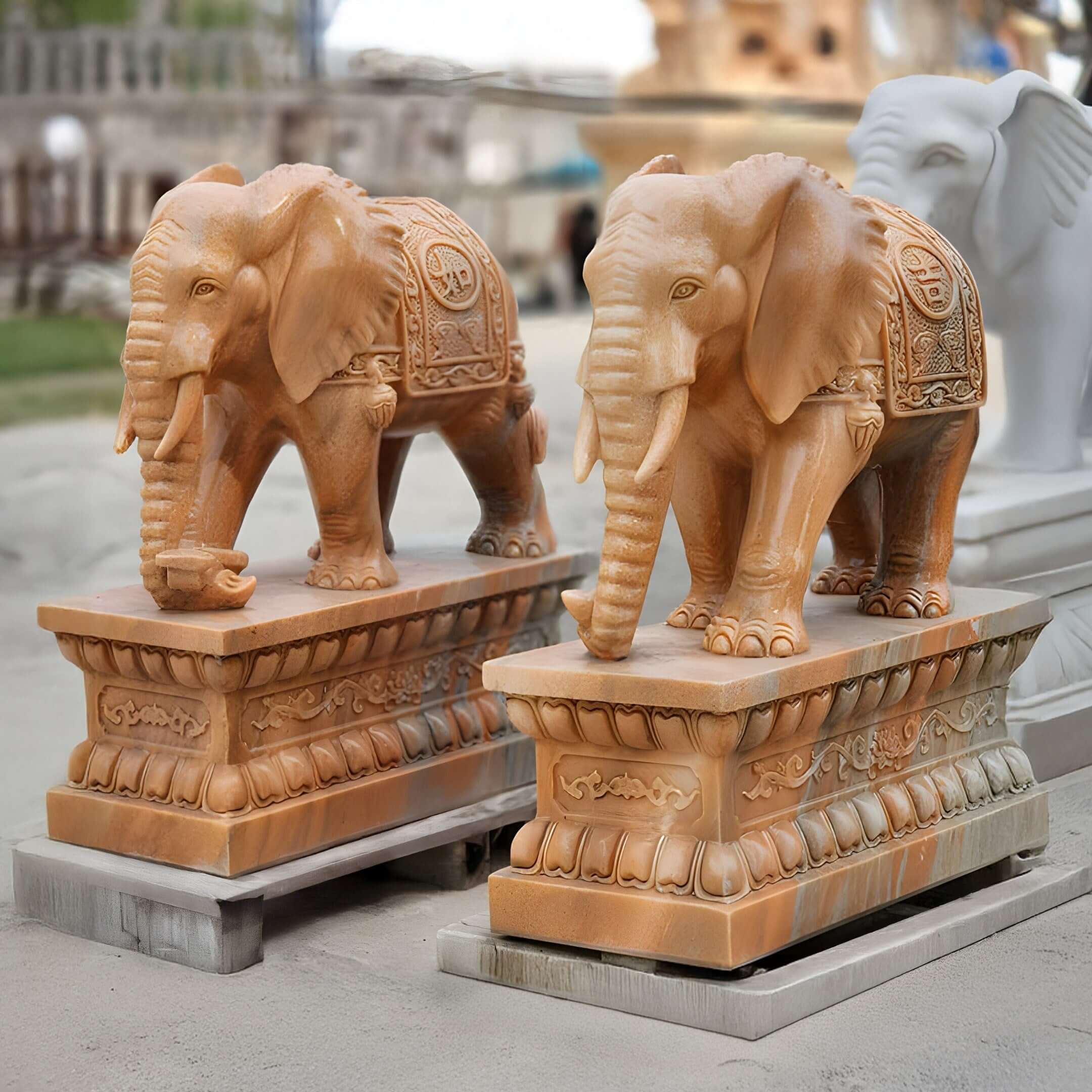Two giant yellow elephant marble sculptures, intricately hand-carved and adorned with ornate designs stand outdoors under a partly cloudy sky. The primary focus is on the exquisite Elephant Yellow Marble Outdoor Sculpture - 180cm by Giant Sculptures, with a third blurred statue in the background.
