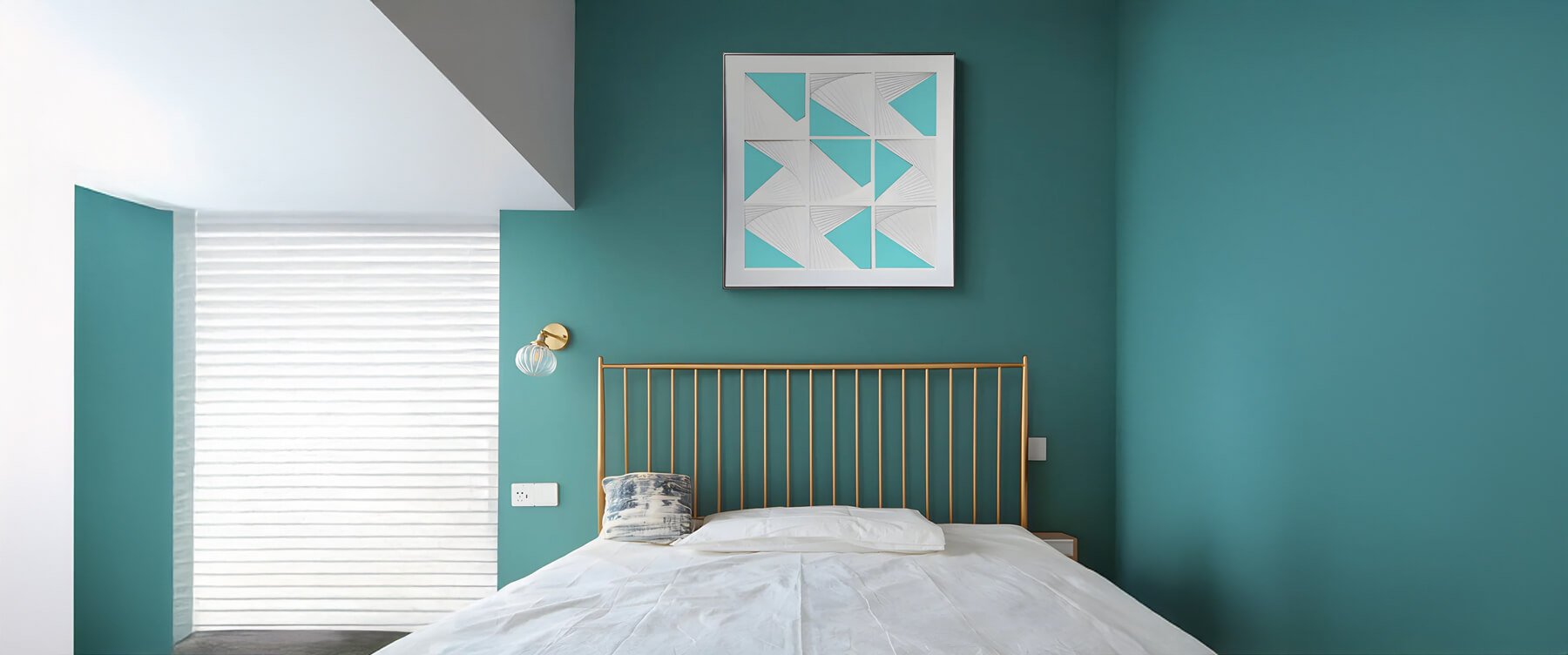 A modern bedroom with teal walls, a minimalist bed with a gold metal headboard, and a white and blue geometric artwork framed above the bed.