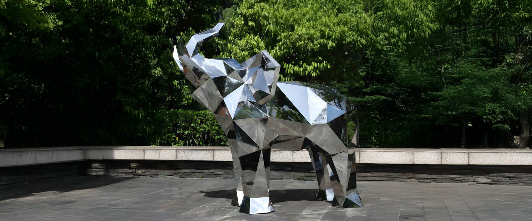 Geometric mirrored elephant sculpture displayed in an outdoor plaza, surrounded by lush green trees and natural light.