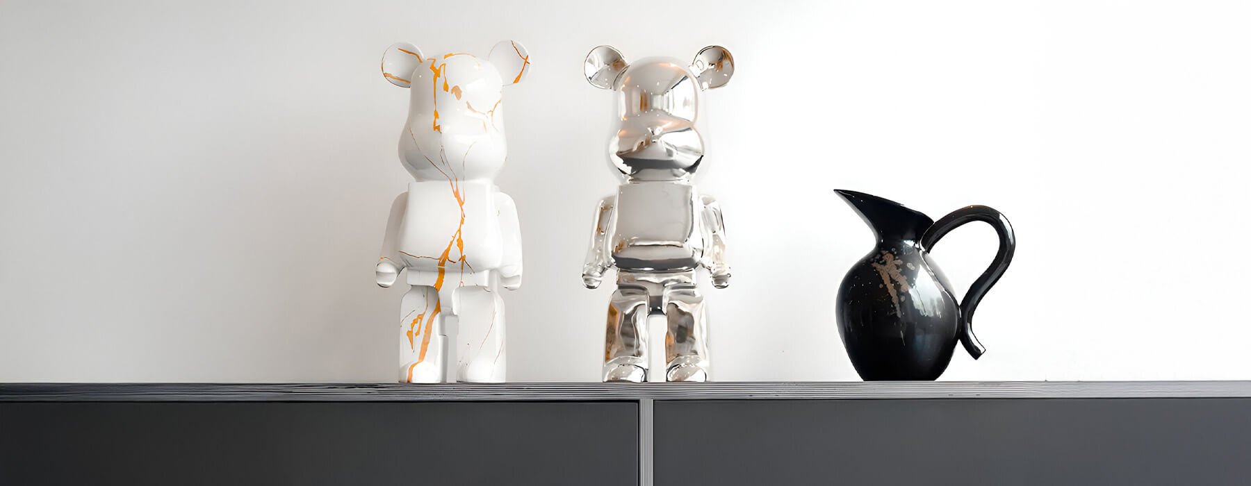 Stylish display featuring white and chrome bear sculptures with gold detailing, accompanied by a sleek black ceramic vase, placed on a modern grey cabinet against a white wall.