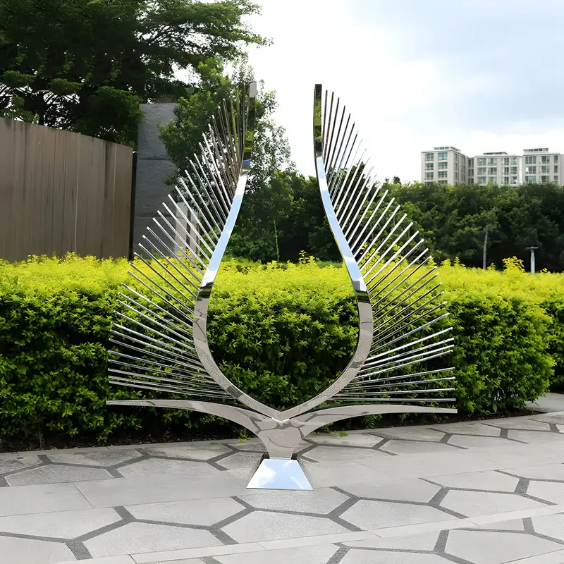 A striking metal sculpture resembling a pair of curved, radiating wings, standing in an outdoor urban setting with green hedges and buildings in the background.